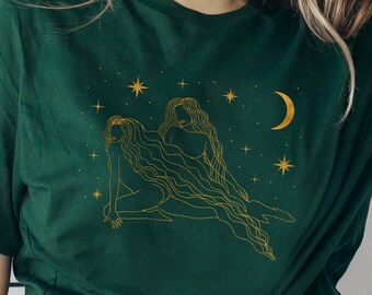 Puede incluir: Camiseta verde oscuro con una ilustración dorada de dos figuras, estrellas y una luna creciente. El diseño está centrado en la camiseta. La camiseta está hecha de un material suave.