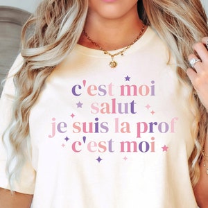 May include: A white t-shirt with the text "c'est moi salut je suis la prof c'est moi" in pink and purple lettering with stars.