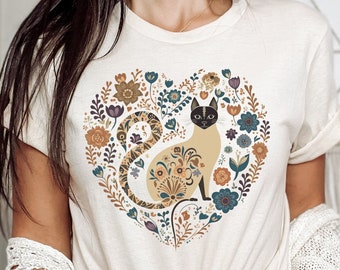 May include: A cream-colored t-shirt with a floral heart design featuring a Siamese cat. The cat is sitting inside the heart and has a black tail and black ears.