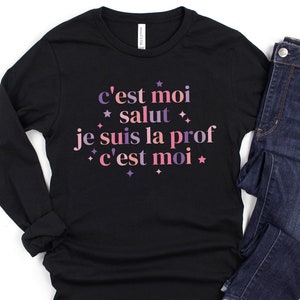 May include: Black long-sleeved shirt with the text "c'est moi salut je suis la prof c'est moi" in pink and purple lettering with stars.