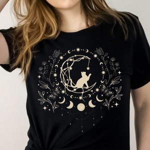 May include: Black t-shirt with a white graphic of a cat sitting on a crescent moon surrounded by stars, flowers, and moons.
