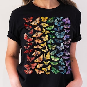 Puede incluir: Una camiseta negra con un estampado de arcoíris de mariposas en la parte delantera. Las mariposas están dispuestas en un degradado de rojo a morado.