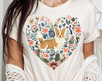 May include: A cream-colored t-shirt with a floral heart design featuring a corgi dog. The heart is filled with colorful flowers and leaves.