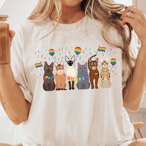 May include: A white t-shirt featuring a graphic of five cats wearing rainbow accessories and holding rainbow flags. The cats are wearing sunglasses and are surrounded by confetti and balloons.