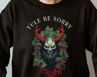 Könnte beinhalten: Ein schwarzes Sweatshirt mit einem grafischen Aufdruck eines gehörnten, grünhäutigen Wesens mit roten Augen und Bart. Das Wesen ist von Stechpalmen und Beeren umgeben. Der Text "YULE BE SORRY" ist über dem Wesen gedruckt.