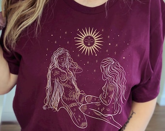 Puede incluir: Una camiseta color burdeos con un diseño gráfico dorado de dos mujeres con cabello largo y detalles intrincados. El diseño incluye un sol con cara y estrellas. El texto "Live away" es visible en el brazo izquierdo de la modelo que lleva la camiseta.