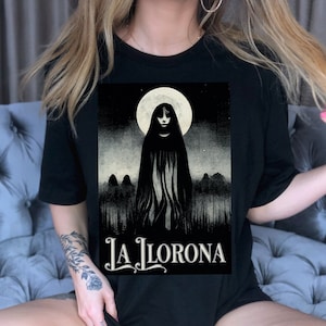 K&ouml;nnte beinhalten: Ein schwarzes T-Shirt mit einem grafischen Aufdruck einer Frau in einem wei&szlig;en Kleid, die vor einem Vollmond steht. Der Text "La Llorona" ist unter dem Grafikdruck aufgedruckt.
