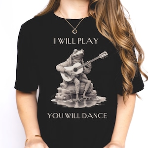 May include: Black t-shirt with a graphic of a frog playing a guitar. The text "I will play, you will dance" is printed on the shirt.