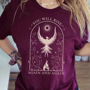 May include: A maroon t-shirt with a white graphic of a phoenix rising from flames inside a floral arch. The text "You Will Rise Again and Again" is printed below the graphic.