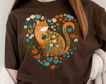 Puede incluir: Una camiseta marrón con un gráfico de un gato sentado en un arreglo de flores en forma de corazón. El gato es naranja con marcas blancas y las flores son en tonos de azul, verde, amarillo y naranja.