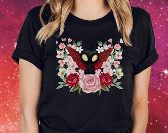 Puede incluir: Camiseta negra con un gráfico de una mariposa roja y negra con ojos brillantes rodeada de flores rosas y rojas.