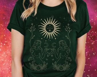 Puede incluir: Una camiseta verde oscuro con un gráfico dorado de dos figuras que se toman de la mano con un sol y una luna sobre ellas. El gráfico está rodeado de estrellas y flores.