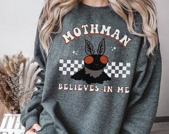May include: A dark gray sweatshirt with a graphic of a moth with red eyes and the text "Mothman Believes in Me" in a retro font.