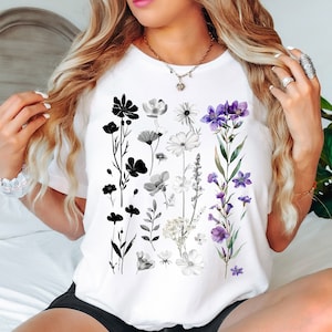 Könnte beinhalten: Ein weißes T-Shirt mit einem floralen Design mit schwarzen und lila Blumen. Die Blumen sind vertikal auf dem Shirt angeordnet.
