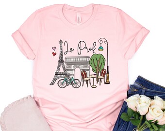May include: Pink t-shirt with a graphic of the Eiffel Tower, the Arc de Triomphe, a bicycle, a street lamp, and a cafe table with chairs. The text "La Prof" is printed on the shirt.