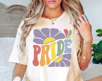 Puede incluir: Camiseta color crema con un diseño floral retro y la palabra "PRIDE" en letras coloridas y onduladas. El diseño presenta una flor morada con un centro amarillo y acentos de arcoíris. La camiseta es de estilo casual y cómodo.