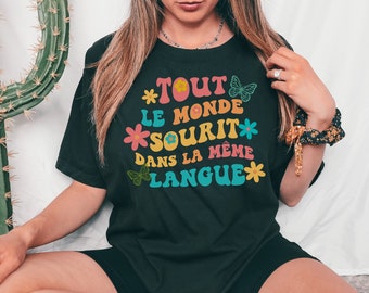 Può includere: T-shirt nero con la scritta francese "Tout le monde sourit dans la même langue" in un font retrò colorato. Il design include fiori e farfalle. La maglietta è in stile casual.