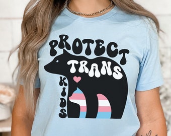 May include: Light blue t-shirt with a black bear and a cat graphic. The text "Protect Trans Kids" is written in a retro font.