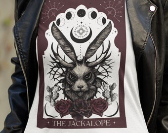 May include: White t-shirt featuring a detailed illustration of a jackalope with antlers, roses, and cacti, framed by a burgundy arch and celestial elements. The text "THE JACKALOPE" is at the bottom. The shirt is worn with denim shorts and a black leather jacket.