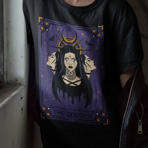 May include: A black t-shirt with a purple tarot card design featuring a woman with three faces and the text "THE GODDESS". The person wearing the shirt has green hair and is wearing a red and black bucket hat.