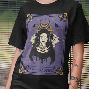 May include: A black t-shirt with a purple and gold illustration of a tarot card. The card is labeled "The Goddess" and features a woman with three faces.