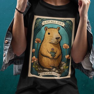 May include: Black t-shirt with a graphic of a capybara holding a flower. The graphic is surrounded by a border with the text "Slay All Day The Capybara".