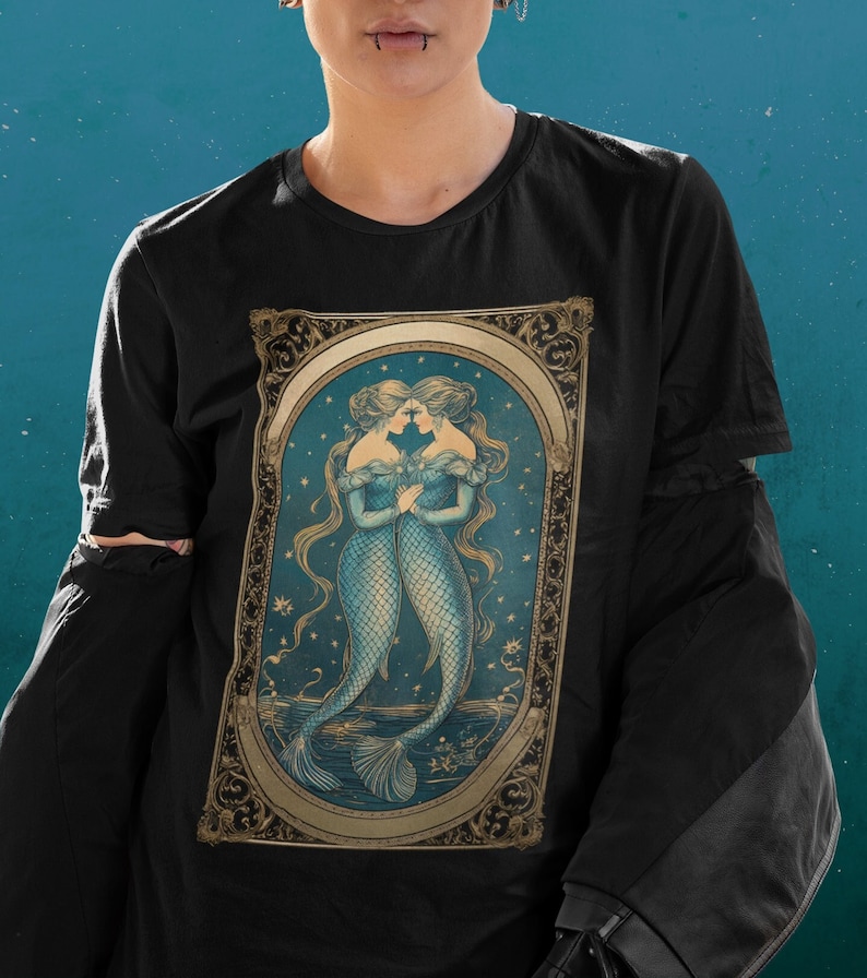 Puede incluir: Camiseta negra con una ilustraci&oacute;n vintage de dos sirenas con colas azules y detalles dorados. Las sirenas se miran y sus colas est&aacute;n entrelazadas. La ilustraci&oacute;n est&aacute; rodeada de un marco dorado con detalles ornamentados.
