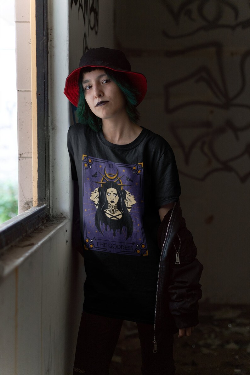 May include: A black t-shirt with a purple tarot card design featuring a woman with three faces and the text "THE GODDESS". The person wearing the shirt has green hair and is wearing a red and black bucket hat.