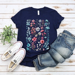 May include: A navy blue t-shirt with a floral print featuring pink, blue, and white flowers. The shirt is laid out on a white wooden surface with a pair of white sneakers and blue jeans.