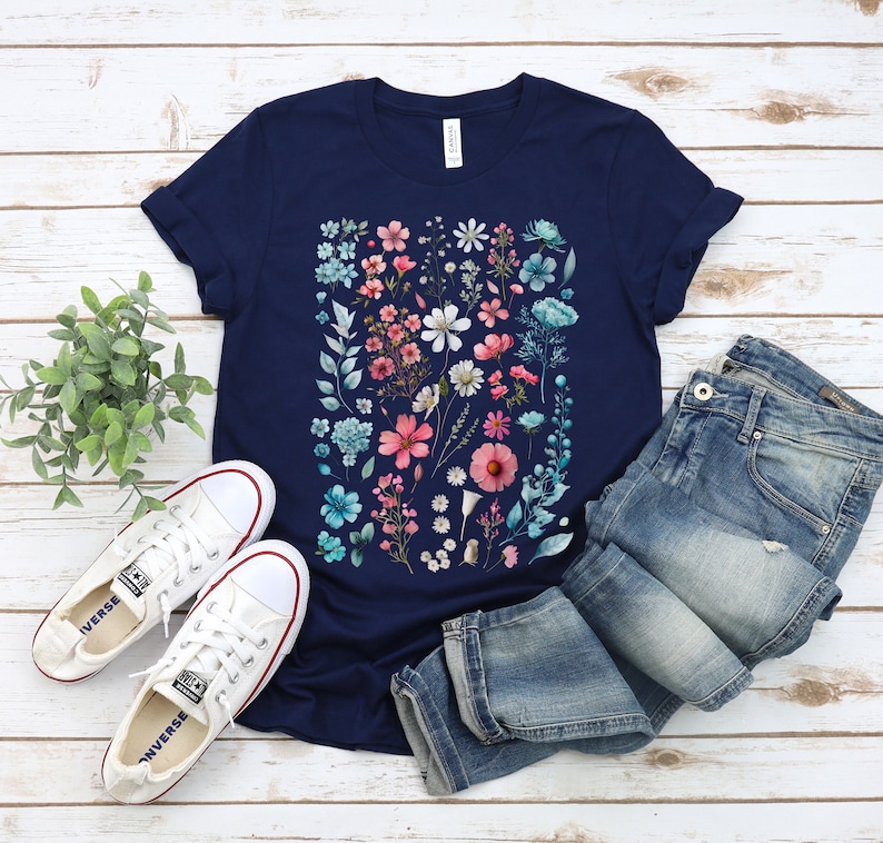 May include: A navy blue t-shirt with a floral print featuring pink, blue, and white flowers. The shirt is laid out on a white wooden surface with a pair of white sneakers and blue jeans.