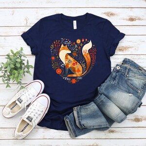 May include: A navy blue t-shirt with a graphic of a fox sitting in a heart-shaped floral design. The fox is orange and white with a floral pattern on its body. The flowers are in shades of orange, red, yellow, and green.