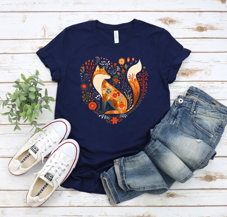May include: A navy blue t-shirt with a graphic of a fox sitting in a heart-shaped floral design. The fox is orange and white with a floral pattern on its body. The flowers are in shades of orange, red, yellow, and green.