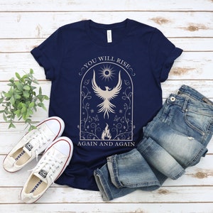 May include: A navy blue t-shirt with a white graphic of a phoenix rising from flames. The graphic is surrounded by a floral design and the text "You Will Rise Again and Again".