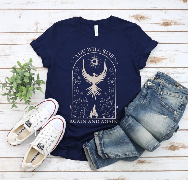 May include: A navy blue t-shirt with a white graphic of a phoenix rising from flames. The graphic is surrounded by a floral design and the text "You Will Rise Again and Again".