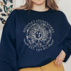Peut inclure: Un sweat-shirt bleu marine avec un imprim&eacute; graphique blanc d'un cercle contenant une t&ecirc;te de M&eacute;duse. Le cercle est entour&eacute; du texte "Medusa's Stoneworks Est. 1350 B.C.".