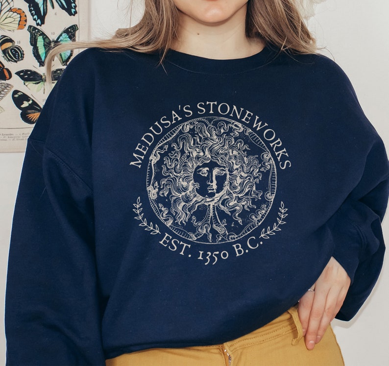 Peut inclure: Un sweat-shirt bleu marine avec un imprim&eacute; graphique blanc d'un cercle contenant une t&ecirc;te de M&eacute;duse. Le cercle est entour&eacute; du texte "Medusa's Stoneworks Est. 1350 B.C.".