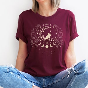 May include: A maroon t-shirt with a gold graphic of a cat sitting on a crescent moon surrounded by stars and floral elements.