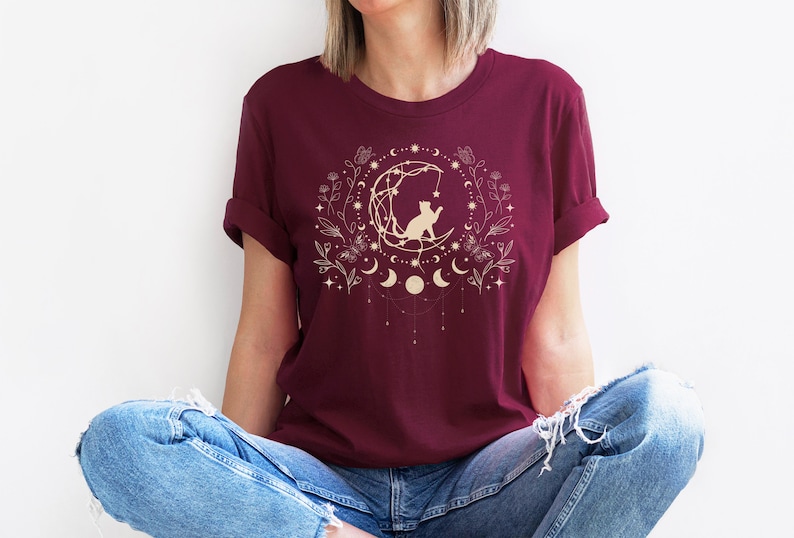 May include: A maroon t-shirt with a gold graphic of a cat sitting on a crescent moon surrounded by stars and floral elements.