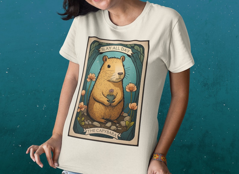 May include: A white t-shirt with a graphic of a cartoon capybara holding a flower. The graphic is surrounded by a blue and green border and the text "SLAY ALL DAY THE CAPYBARA".