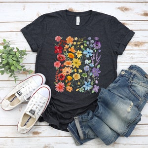 Puede incluir: Una camiseta gris oscuro jaspeada con un estampado floral arco&iacute;ris. Las flores son en tonos de rojo, naranja, amarillo, verde, azul y morado. La camiseta es de corte holgado y tiene cuello redondo.