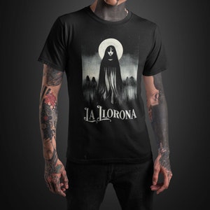 K&ouml;nnte beinhalten: Schwarzes T-Shirt mit einem Grafikdruck einer Frau in einem wei&szlig;en Kapuzengewand, die vor einem Vollmond steht. Der Text "LA LLORONA" ist unter dem Bild gedruckt.