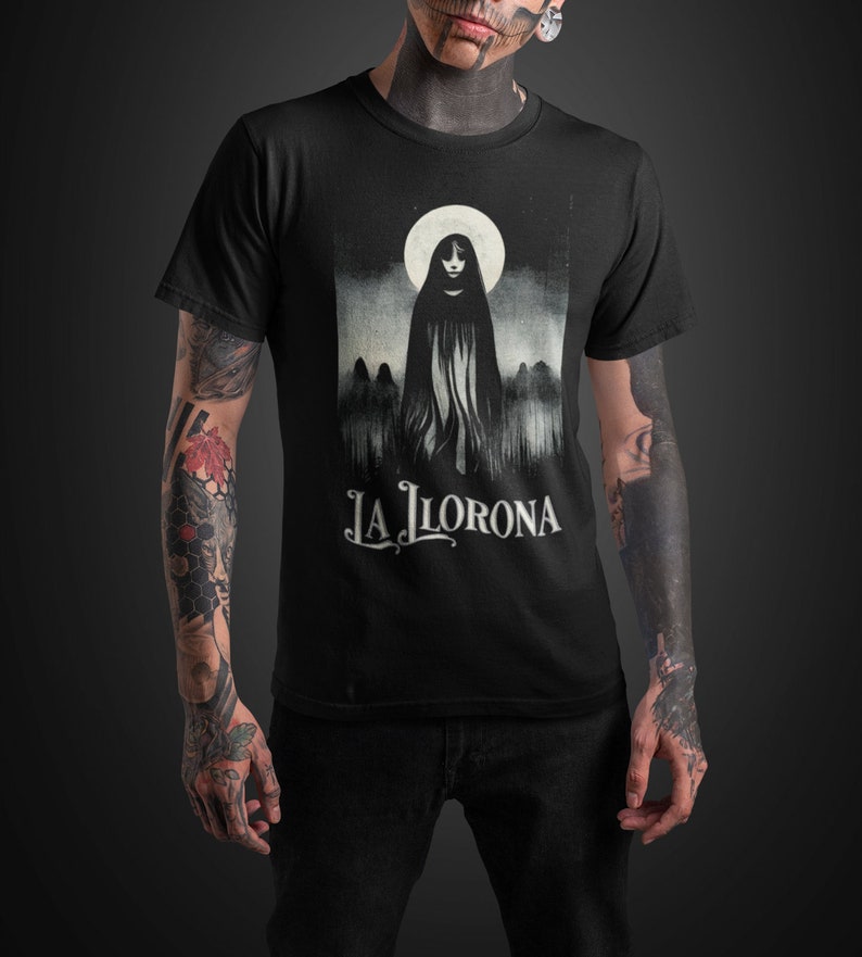 K&ouml;nnte beinhalten: Schwarzes T-Shirt mit einem Grafikdruck einer Frau in einem wei&szlig;en Kapuzengewand, die vor einem Vollmond steht. Der Text "LA LLORONA" ist unter dem Bild gedruckt.