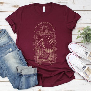 K&ouml;nnte beinhalten: Ein maroonfarbenes T-Shirt mit einem grafischen Design aus einer Bergkette, einem Halbmond, Sternen und einem Buch. Der Text "Fictional Wanderlust" ist auf das Shirt gedruckt.