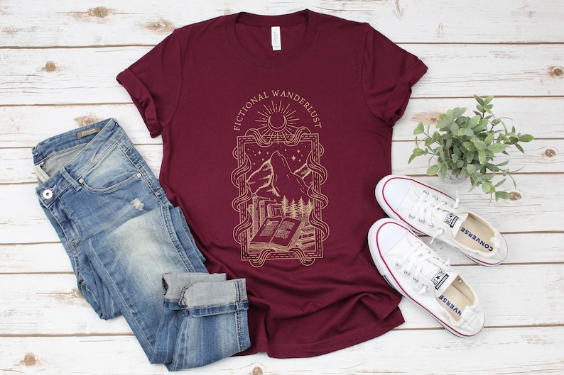 K&ouml;nnte beinhalten: Ein maroonfarbenes T-Shirt mit einem grafischen Design aus einer Bergkette, einem Halbmond, Sternen und einem Buch. Der Text "Fictional Wanderlust" ist auf das Shirt gedruckt.
