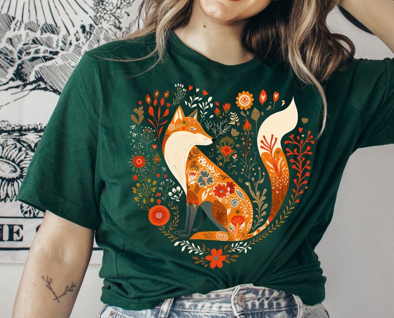 May include: A green t-shirt with a colorful fox illustration surrounded by flowers. The fox is orange, white, and brown with floral patterns. The flowers are red, orange, yellow, and green.