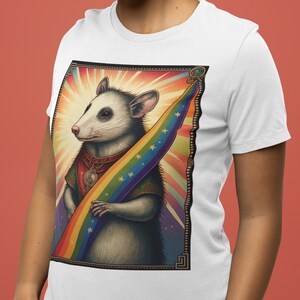 Pode incluir: Camiseta branca com uma ilustra&ccedil;&atilde;o colorida de um gamb&aacute; segurando uma bandeira arco-&iacute;ris. O gamb&aacute; est&aacute; usando um colar com um s&iacute;mbolo yin yang e uma coroa. O fundo &eacute; um design abstrato colorido.