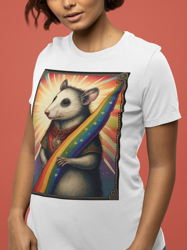 Pode incluir: Camiseta branca com uma ilustra&ccedil;&atilde;o colorida de um gamb&aacute; segurando uma bandeira arco-&iacute;ris. O gamb&aacute; est&aacute; usando um colar com um s&iacute;mbolo yin yang e uma coroa. O fundo &eacute; um design abstrato colorido.
