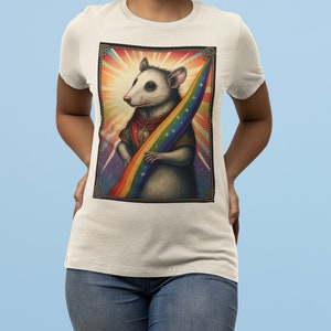 Pode incluir: Uma camiseta branca com um gr&aacute;fico de um gamb&aacute; segurando uma bandeira arco-&iacute;ris. O gamb&aacute; est&aacute; usando um colar com um pingente de caveira. O fundo &eacute; um design abstrato e colorido.