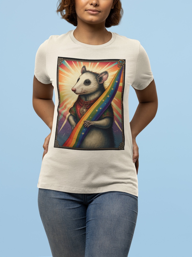 Pode incluir: Uma camiseta branca com um gr&aacute;fico de um gamb&aacute; segurando uma bandeira arco-&iacute;ris. O gamb&aacute; est&aacute; usando um colar com um pingente de caveira. O fundo &eacute; um design abstrato e colorido.