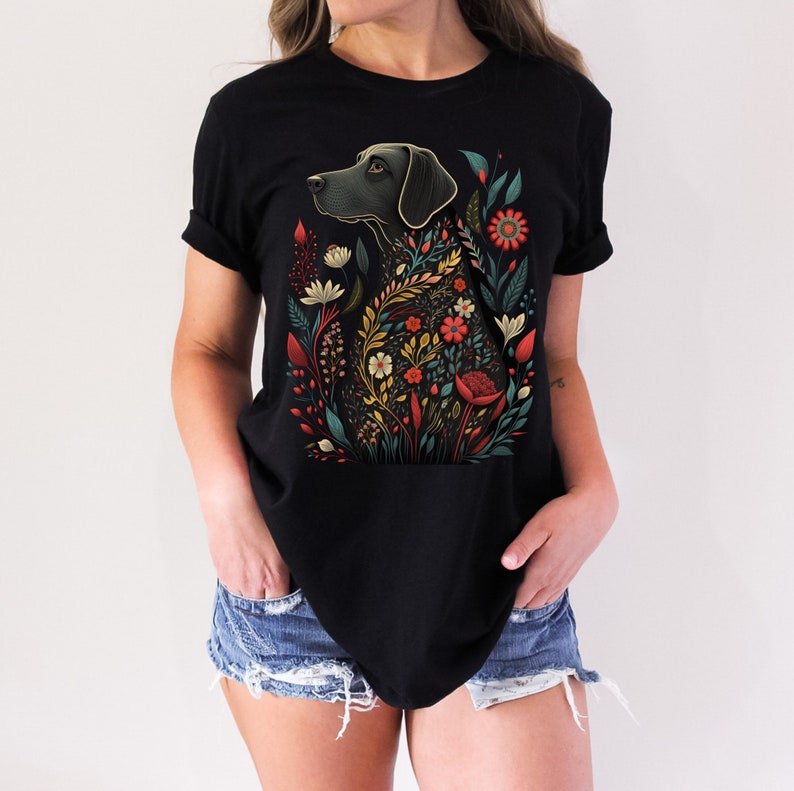 May include: Black t-shirt with a floral design featuring a black dog. The dog is surrounded by colorful flowers and leaves.